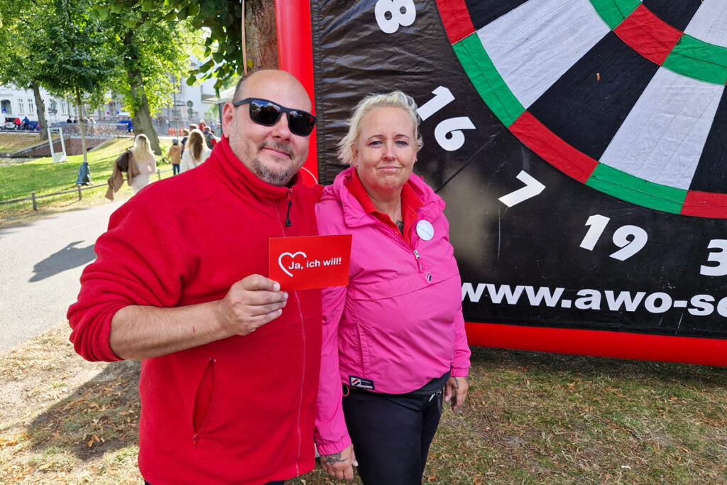 Zwei Personen stehen l&auml;chelnd vor einem gro&szlig;en Dartboard.
