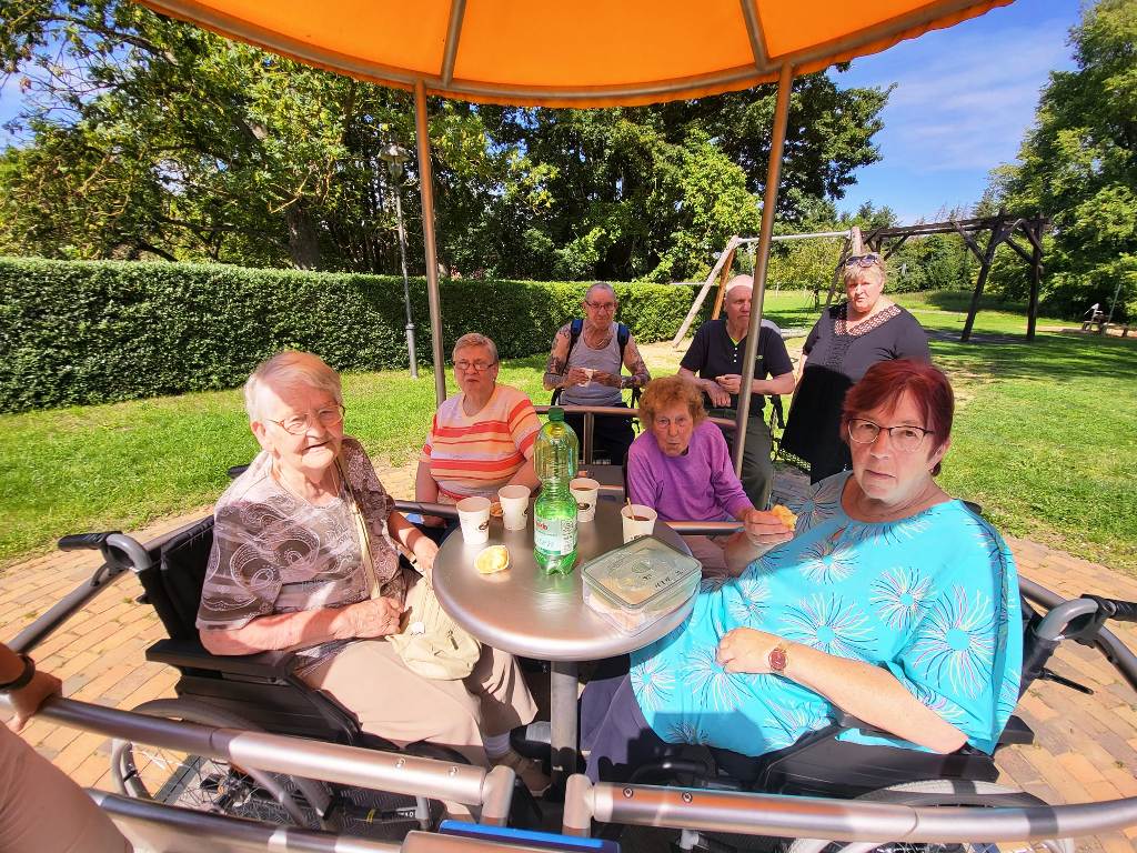 Eine Gruppe älterer Menschen sitzt im Freien an einem Tisch unter einem orangen Sonnenschirm.