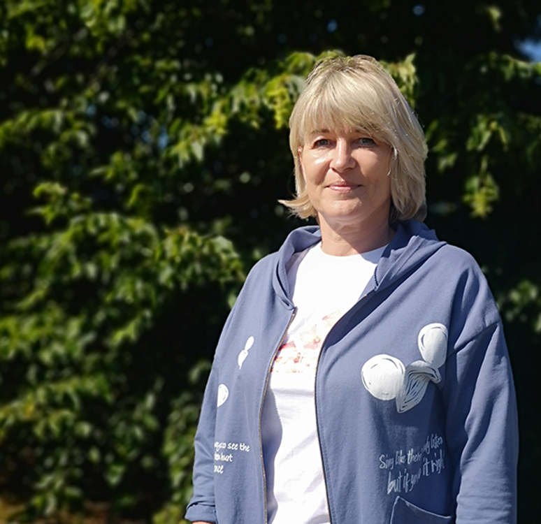 Frau in blauer Kapuzenjacke steht im Freien vor einem Hintergrund aus grünen Bäumen.