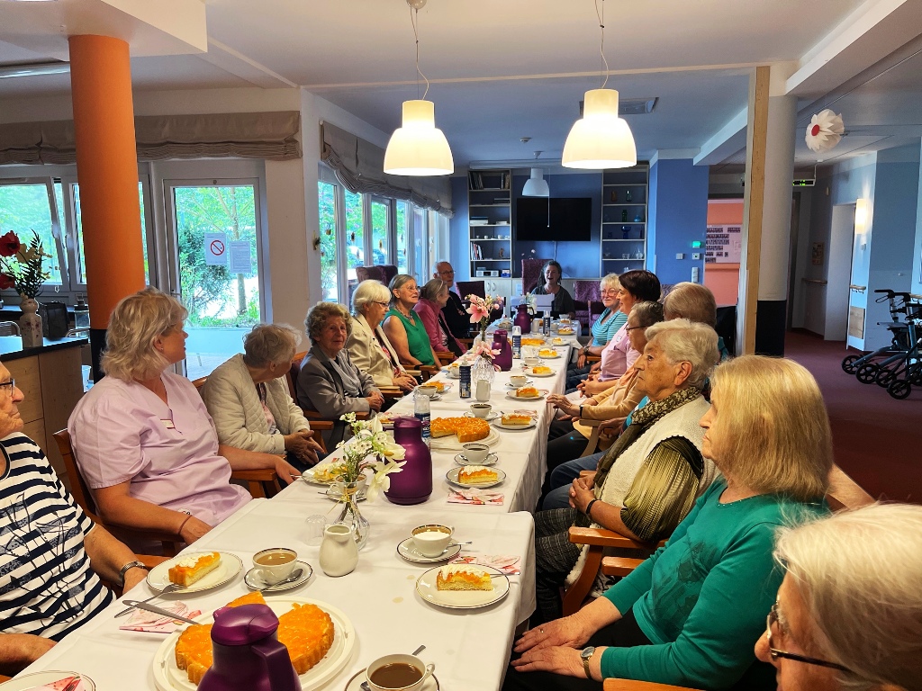 Personen sitzen an einem langen Tisch, gedeckt mit Kuchen und Getränken.
