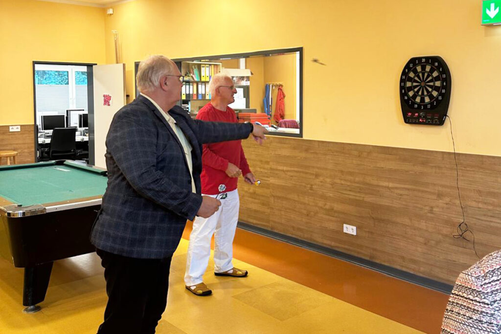 Zwei M&auml;nner spielen Dart in einem Raum mit Billardtisch