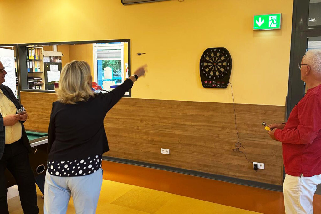 Drei Personen spielen Dart an einer Dartscheibe in einem Raum.