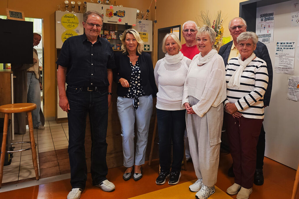 Gruppe von Menschen in einem Raum mit hellen W&auml;nden, Regalen und Plakaten im Hintergrund.