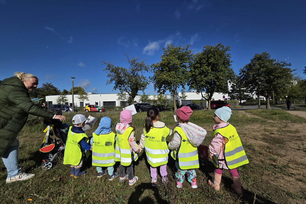 Eine Gruppe von kleinen Kindern in leuchtend gelben Sicherheitswesten h&auml;lt sich im Freien an den H&auml;nden.