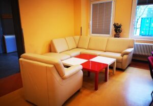 Bild eines kleinen Wohnbereichs mit einer cremefarbenen Eckcouch und einem roten Tisch.