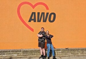 Zwei Personen stehen vor einer Wand mit AWO-Logo.