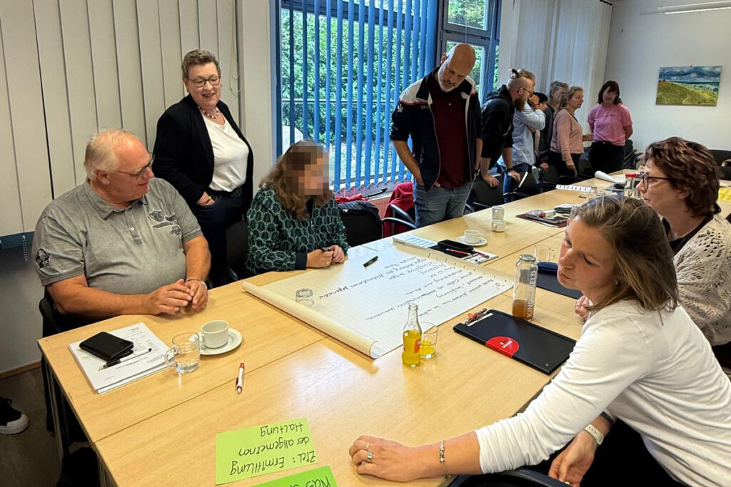 Menschen sitzen und stehen um einen Konferenztisch und diskutieren, mit Notizen auf einem gro&szlig;en Blatt Papier in der Mitte.