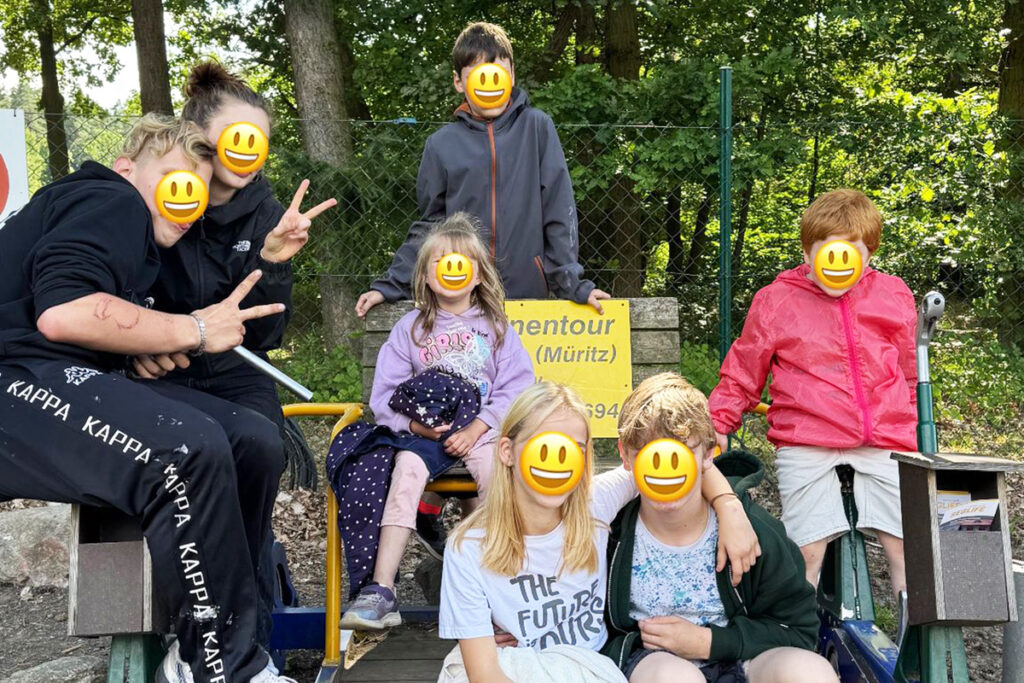 Eine Gruppe von lachenden Kindern und Jugendlichen sitzt und steht auf einem Kettcar im Freien vor einem Schild mit dem Wort 'Draisinentour'.
