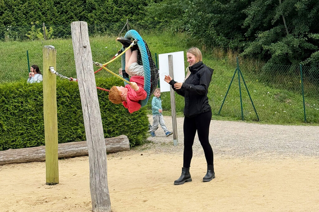 Ein Kind schaukelt in einer Vogelnestschaukel auf einem Spielplatz, w&auml;hrend eine Frau daneben steht und ihm zuschaut.