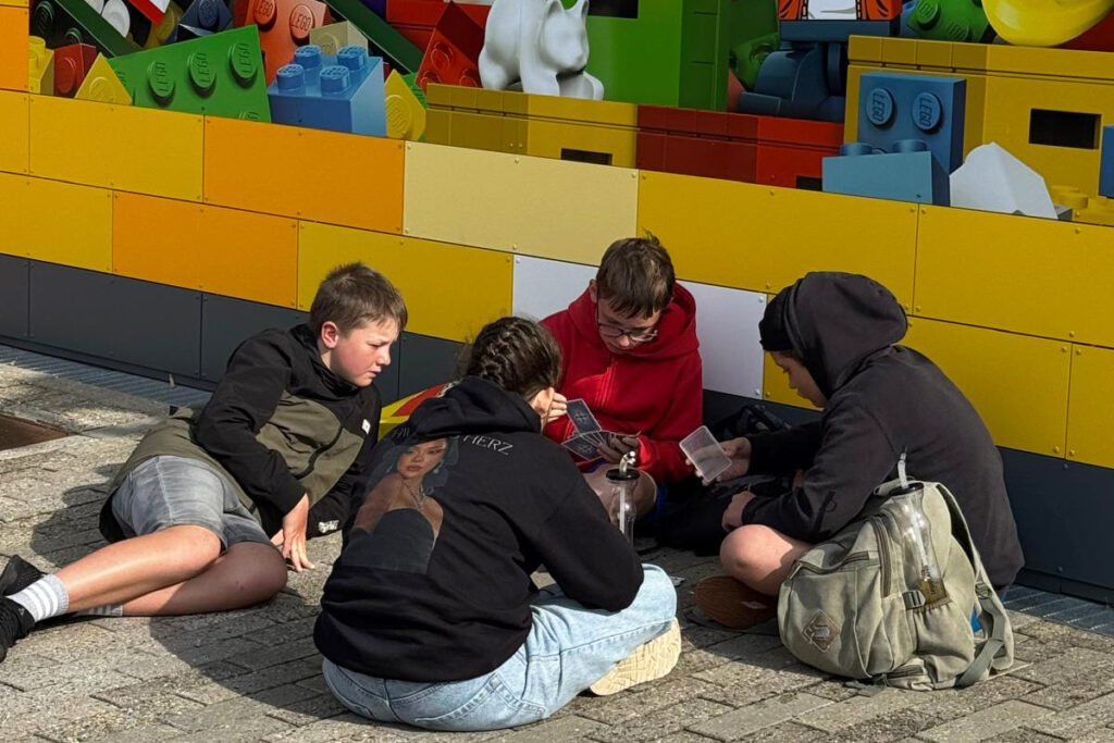 Vier Kinder sitzen auf dem Boden und spielen Karten vor einer bunten Lego-Wand im Freien.
