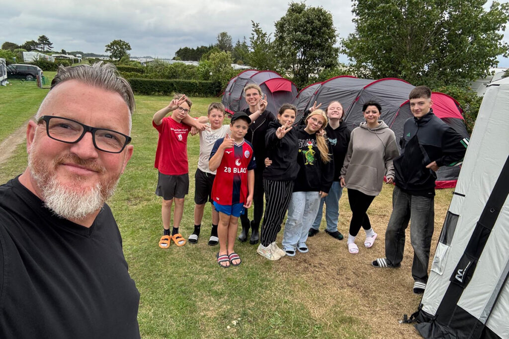 Eine Gruppe von Menschen steht l&auml;chelnd auf einem Campingplatz mit Zelten im Hintergrund.