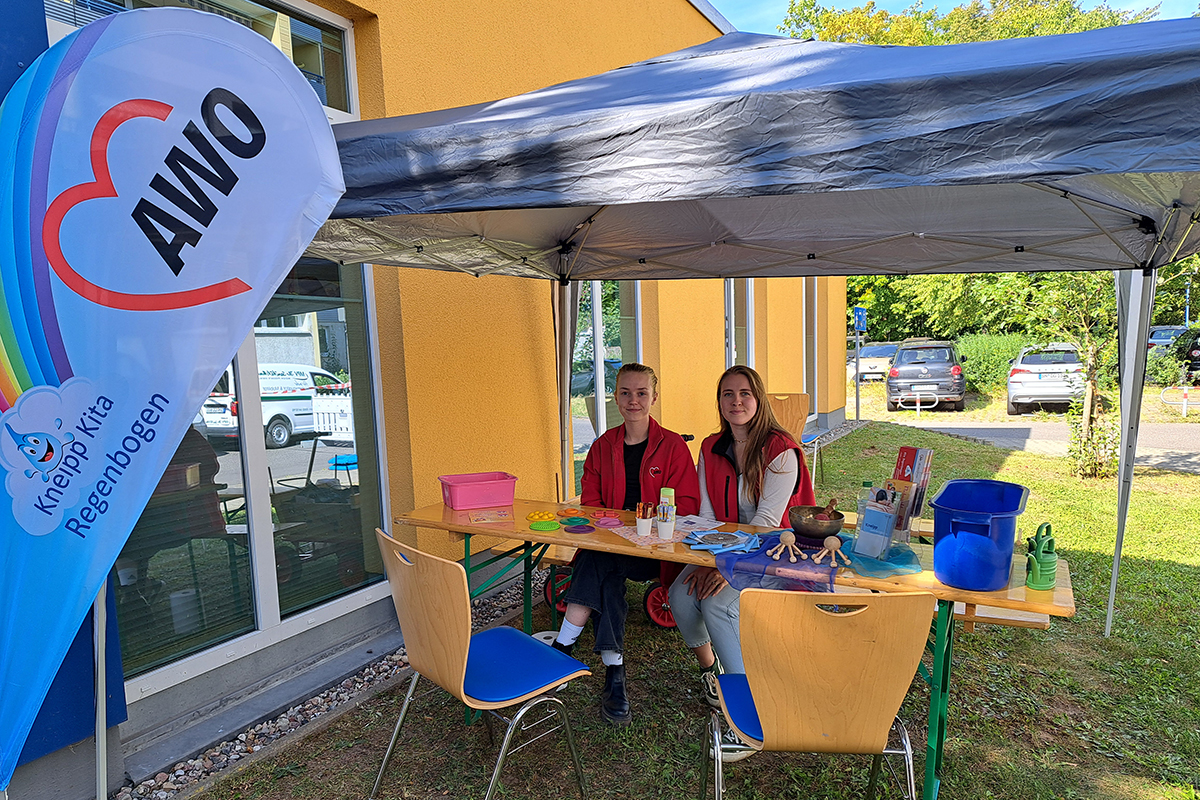 Zwei Personen sitzen an einem Tisch unter einem Pavillon neben einem AWO-Banner.