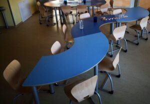 Ein leerer Raum mit blauen Tischen in geschwungener Form und hellen Holzstühlen.