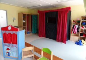 Ein Raum mit einem roten und schwarzen Puppentheater-Vorhang, einem kleinen blauen in Zimmermitte mit einem Clown darauf, Holzregalen mit Verkleidungsstücken und Kindermöbeln.