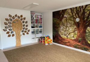 Ein Raum mit einem Baum-Wandbild, Regalen und einem Spielbereich.