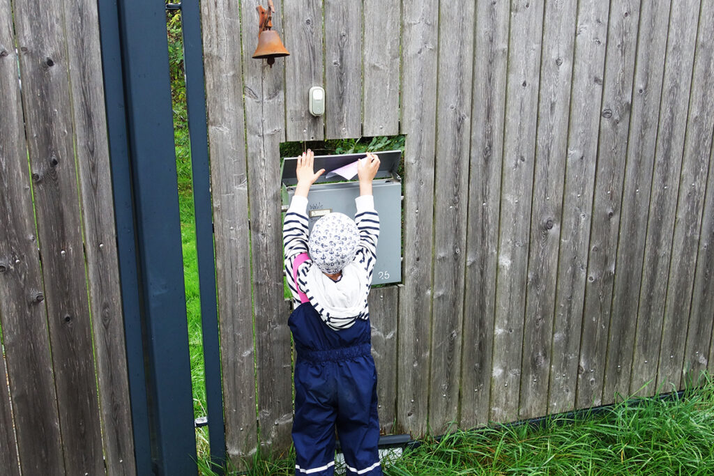 Ein Kind in Regenkleidung steckt einen Brief in einen Briefkasten an einem Holzzaun.