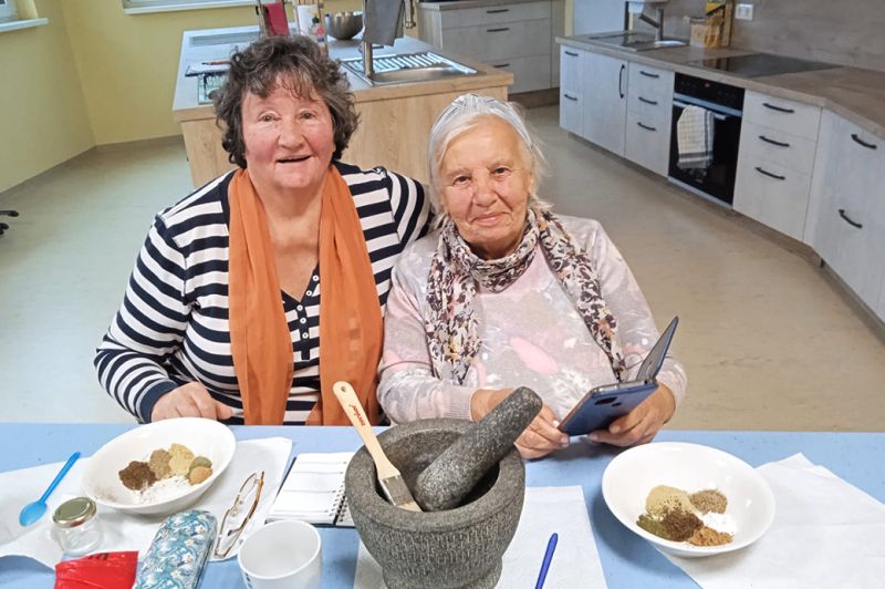 Zwei ältere Frauen sitzen an einem Tisch in einer Küche.