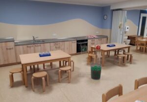 Innenansicht einer Kindertagesstätte mit Holztischen und Stühlen sowie einer Küchenzeile.