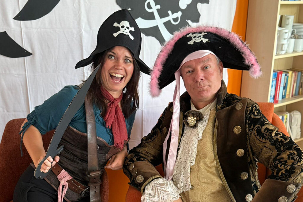 Zwei Personen in Piratenkost&uuml;men vor einem gro&szlig;en Piratenflaggen-Hintergrund.