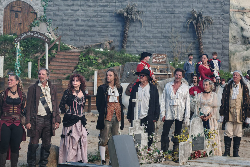 Ensemble von Schauspielern in historischer Kleidung vor einer Kulisse mit Steinmauer und Treppe.