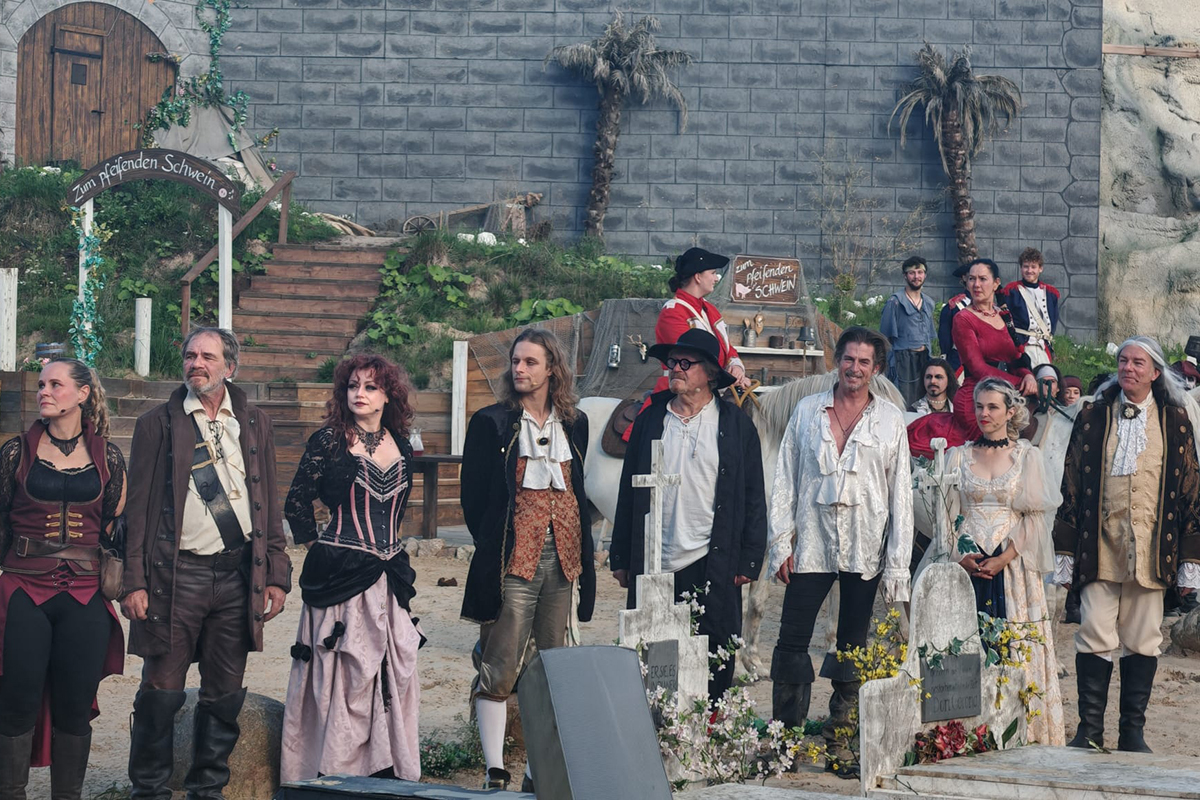 Ensemble von Schauspielern in historischer Kleidung vor einer Kulisse mit Steinmauer und Treppe.