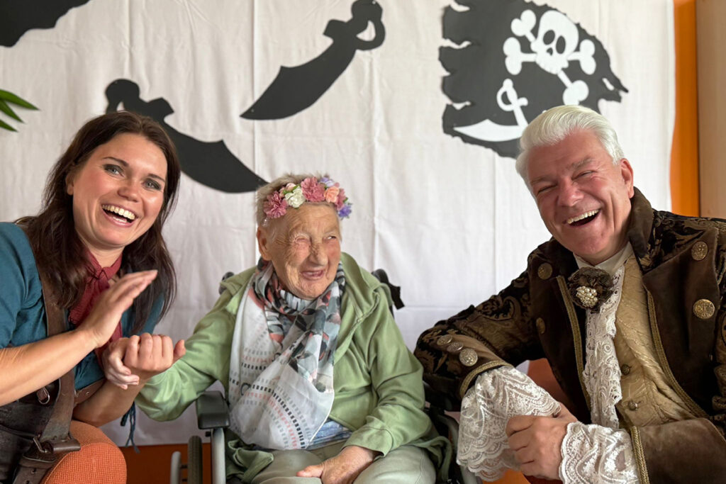 Eine &auml;ltere Frau mit Blumenkranz auf dem Kopf sitzt gl&uuml;cklich in einem Rollstuhl zwischen einem lachenden jungen Mann im historischen Kost&uuml;m und einer lachenden Frau.