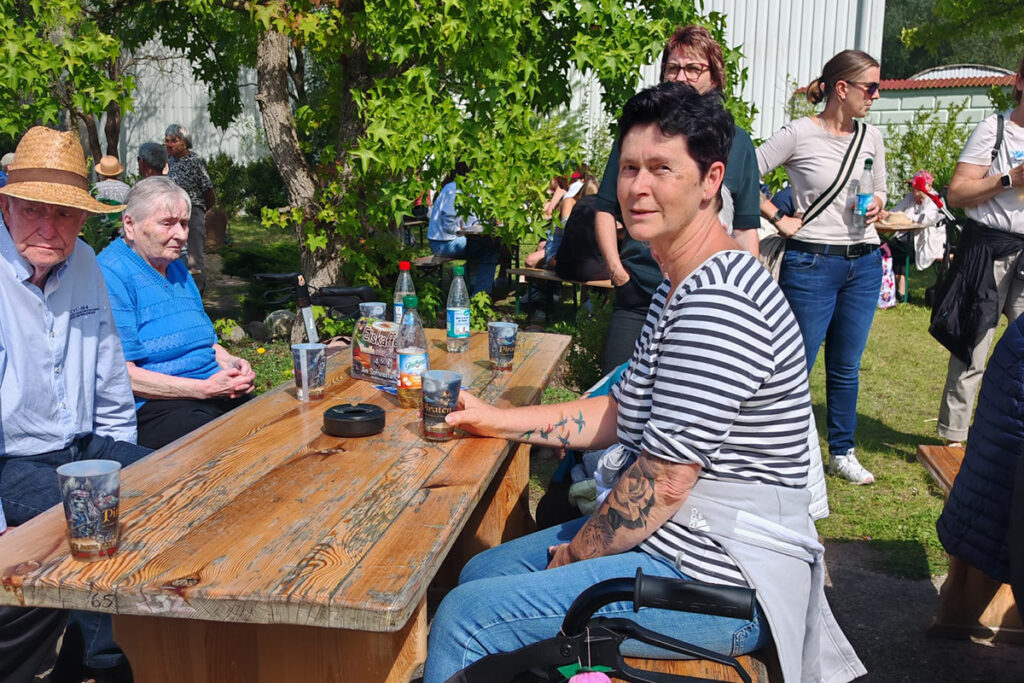 Menschen sitzen und stehen um einen Holztisch im Freien bei sonnigem Wetter.