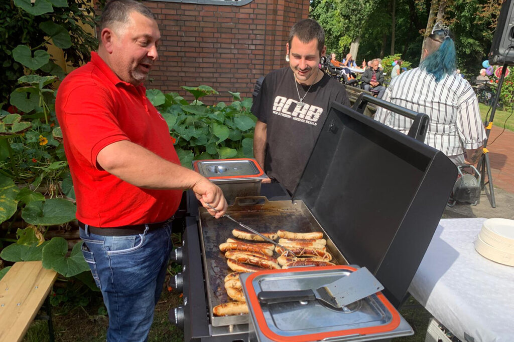 Zwei M&auml;nner beim Grillen im Freien. Einer tr&auml;gt ein rotes Hemd und wendet W&uuml;rste auf einem Grill, der andere schaut zu. Im Hintergrund sind Pflanzen und weitere Personen zu sehen.