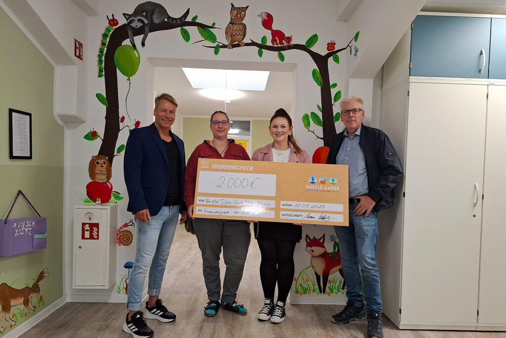 Gruppe von vier Personen mit einem großen Spendenscheck in einem bunt dekorierten Raum.