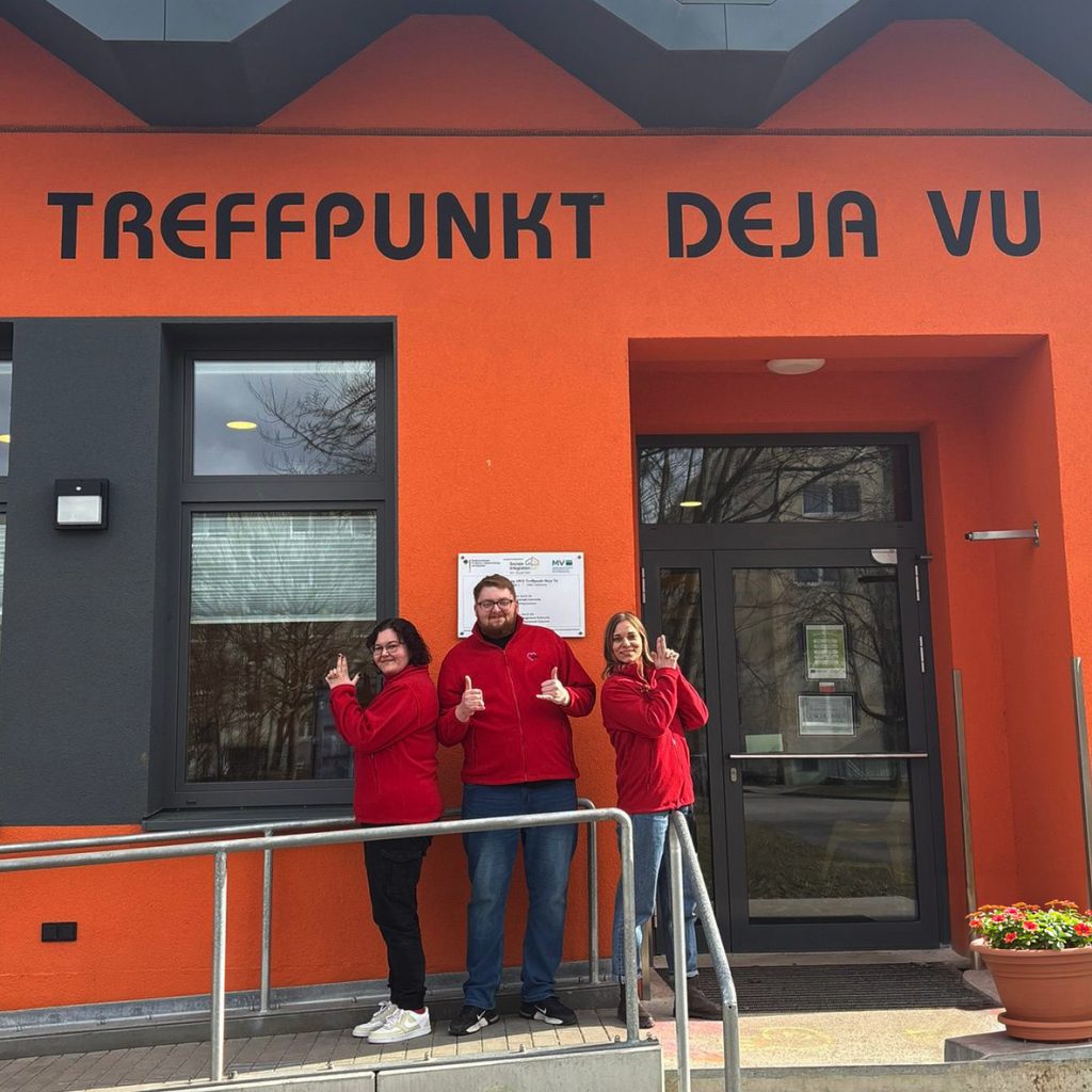 Drei Personen in roten Oberteilen vor dem Eingang des AWO Treffpunkts „Deja Vu“ in einem orangefarbenen Gebäude mit Rampe und Treppe.