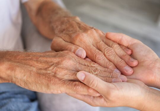 Ein Bild, das zwei paar Hände zeigt, wobei die Hände einer jüngeren Person die Hände einer älteren Person halten.