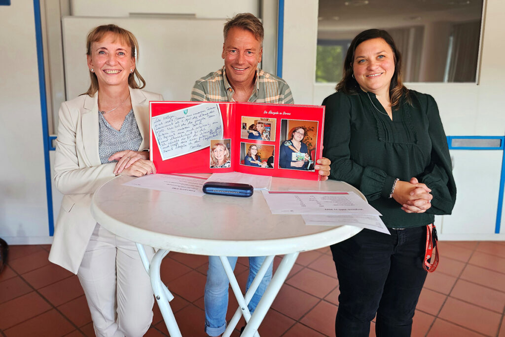 Drei Personen stehen an einem runden Tisch und halten ein rotes Fotoalbum aufgeschlagen in die Kamera.
