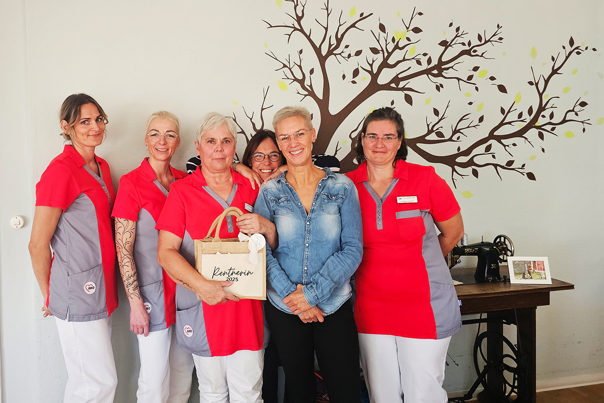 Sechs Frauen in roter und grauer Arbeitskleidung sowie einer in Jeanshemd, lächeln in die Kamera. Hinter ihnen ist eine Wand mit einem gemalten Baum zu sehen.