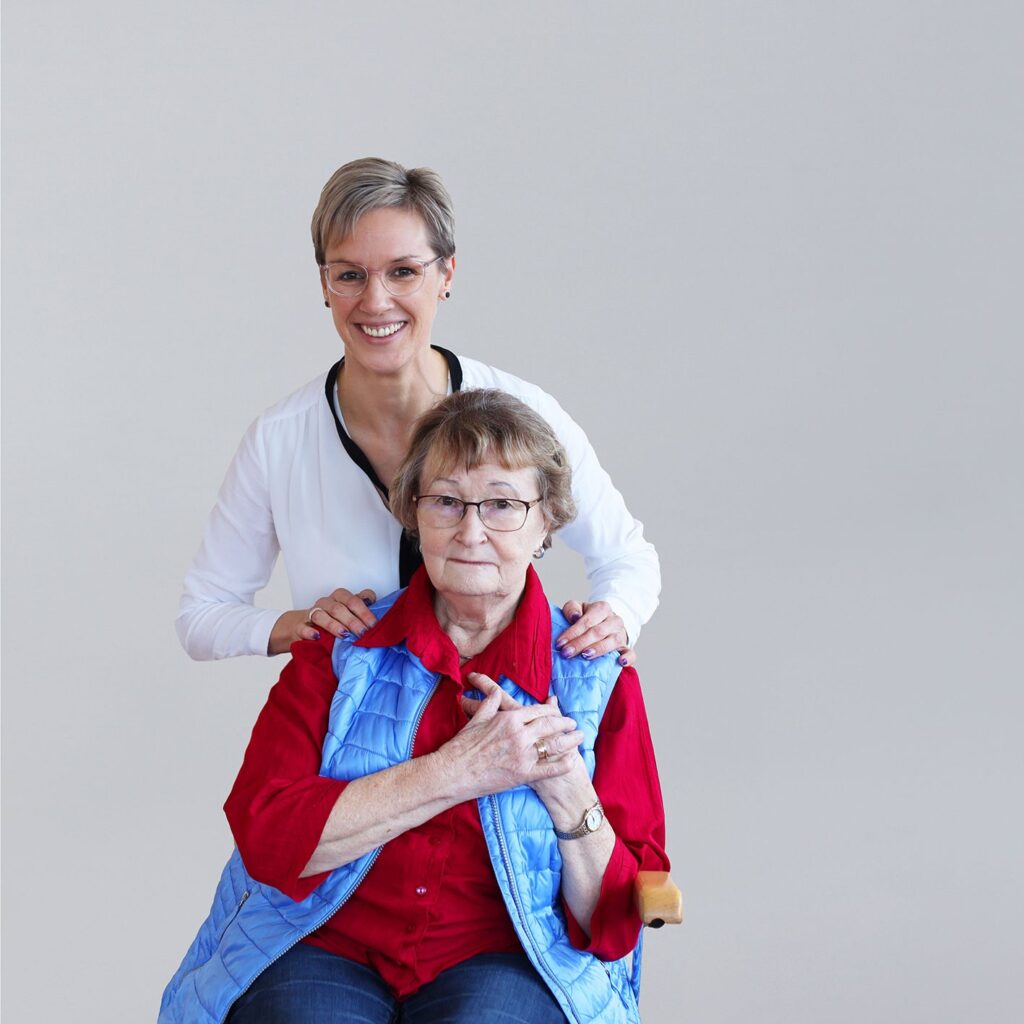 Eine ältere Frau sitzt auf einem Stuhl, hinter ihr steht eine jüngere Frau, beide lächeln.