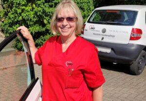 Frau in roter Arbeitskleidung von AWO steht lächelnd neben einem Auto.