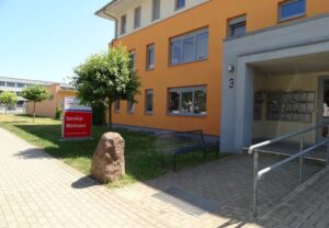 Modernes Gebäude mit orangefarbener Fassade und Schild 'Service Wohnen' davor.