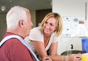 Eine Betreuerin unterhält sich freundlich mit einem älteren Mann in einem Pflege- oder Betreuungsumfeld.