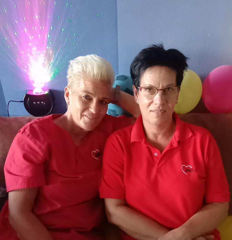 Zwei Frauen in roten Arbeitsuniformen sitzen eng beieinander auf einem Sofa.