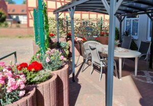 Ein überdachter Außenbereich mit einem Tisch und Stühlen umgeben von Blumenkästen.
