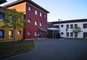 Zweistöckiges Gebäude in rot-gelber Farbe mit mehreren Fenstern und einem grünen Baum davor.