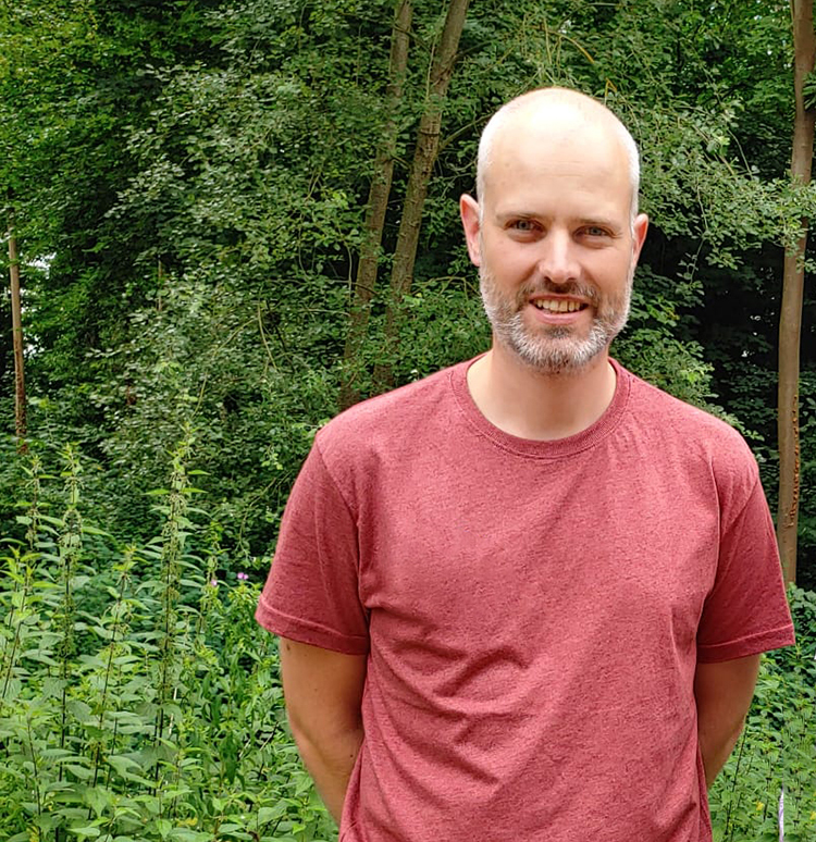 Person in einem roten T-Shirt lächelt in die Kamera, vor einem Hintergrund aus Bäumen und dichtem Laubwerk.