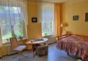 Innenansicht eines gemütlichen Wohnzimmers oder Pflegezimmers mit einem Bett, Tisch und Stühlen.