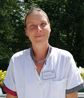 Eine Frau in weißem medizinischen Kittel mit Namensschild, vor einem Hintergrund aus Bäumen.