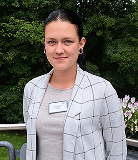 Eine Frau mit dunklem, zurückgebundenem Haar steht draußen vor einer grünen Kulisse. Sie trägt einen karierten Blazer und ein Namensschild.