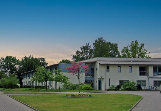 Ansicht eines modernen Gebäudekomplexes mit Vordach und umliegender Grünfläche bei klarem Himmel.