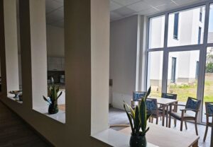 Innenaufnahme eines modernen Gemeinschaftsraums mit großen Fenstern, Pflanzen und Sitzgelegenheiten.
