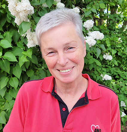 Person with short gray hair wearing a red AWO polo shirt standing in front of green foliage and white flowers.