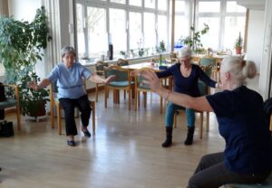 Drei ältere Frauen machen gymnastische Übungen im Sitzen in einem Gemeinschaftsraum.