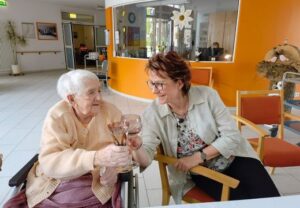 Zwei Frauen sitzen zusammen und stoßen in einem hellen Raum mit Glasgefäßen an.