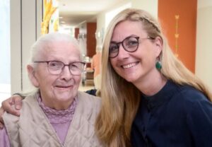 Eine ältere Frau und eine jüngere Frau stehen in einem Flur und lächeln in die Kamera.